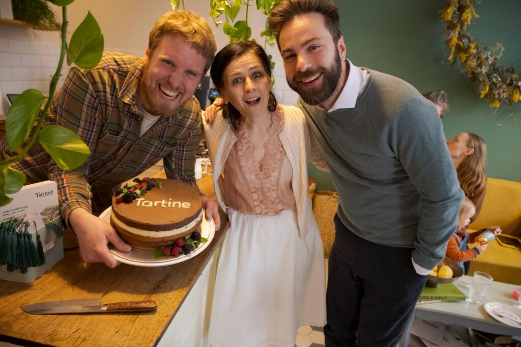 Das Team des Café Tartine: Drei lachende Personen posieren vor dem Tresen. Der linke hält eine Torte hoch, auf der in weißem Zuckerguss der Schriftzug "Tartine" steht