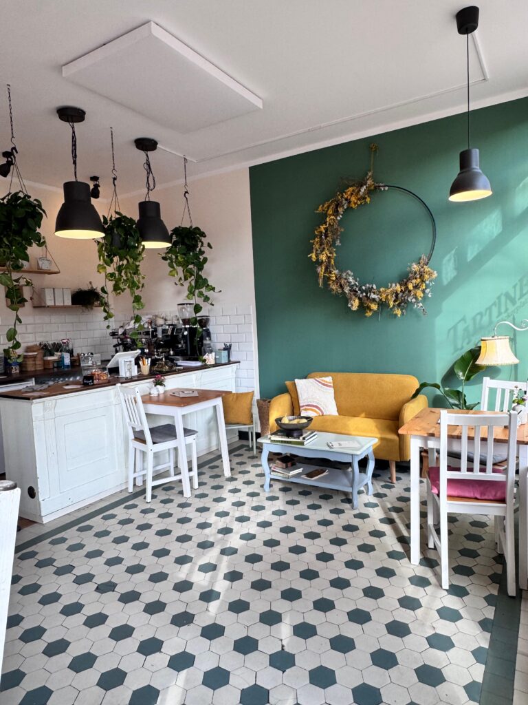 Innenansicht des Café Tartine, das eine Mischung aus modernem und gemütlichem Stil zeigt, mit grün gestrichenen Wänden, hellen Holzmöbeln und Fliesenboden. Eine gelbe Couch steht in einer Ecke, davor ein hellblauer Couchtisch.